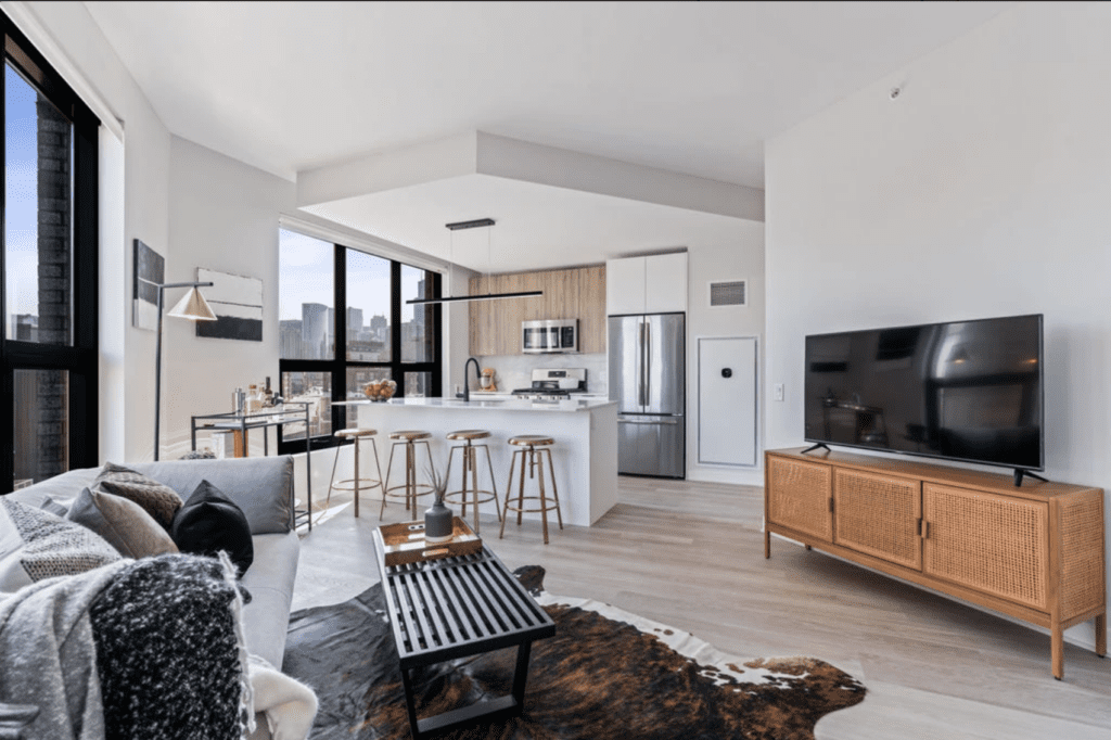 Modern living room with an open kitchen, featuring a large TV on a wooden stand, a white island with stools, a city view from large windows, and a cowhide rug on light wood flooring.