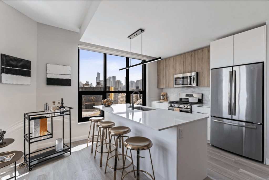 Modern kitchen with white cabinets, island with stools, stainless steel appliances, and large window showing cityscape. Art and a bar cart adorn the space. Light wood flooring is present.