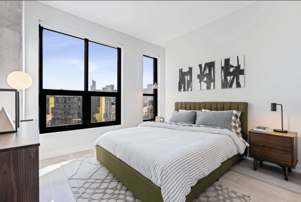 A modern bedroom with a green upholstered bed, striped bedding, two wooden nightstands, abstract wall art, and large windows showing a city view.