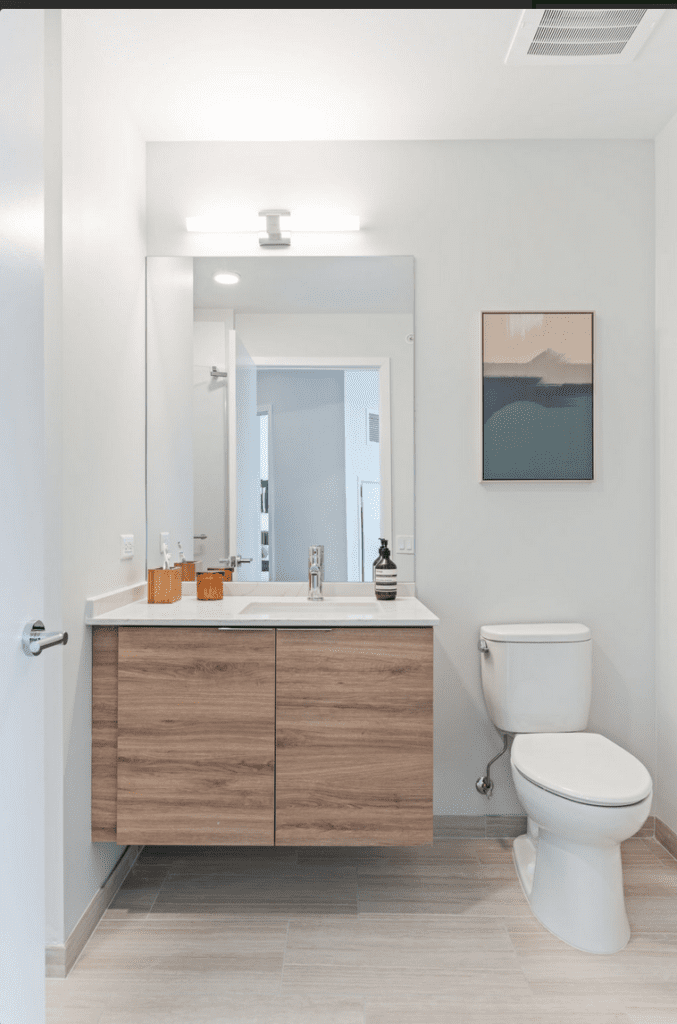 Modern bathroom with a wood cabinet, white countertop, sink, and mirror. Toilet on the right, abstract art on the wall, and beige floor tiles.