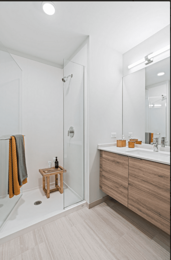 Modern bathroom with glass shower, wooden stool, and towels. Vanity with dual sinks, large mirror, and wooden cabinets. Bright lighting and neutral decor.