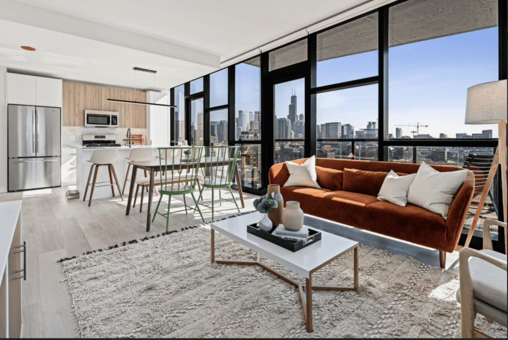 Modern living room and kitchen with floor-to-ceiling windows. Features a brown sofa, white coffee table, and bar stools, overlooking a city skyline.