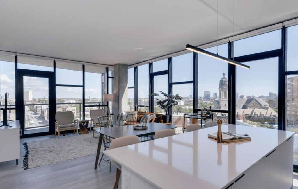 Modern open-concept living space with large windows, city view, white kitchen island, dining area, and seating arrangement. Natural light fills the room.