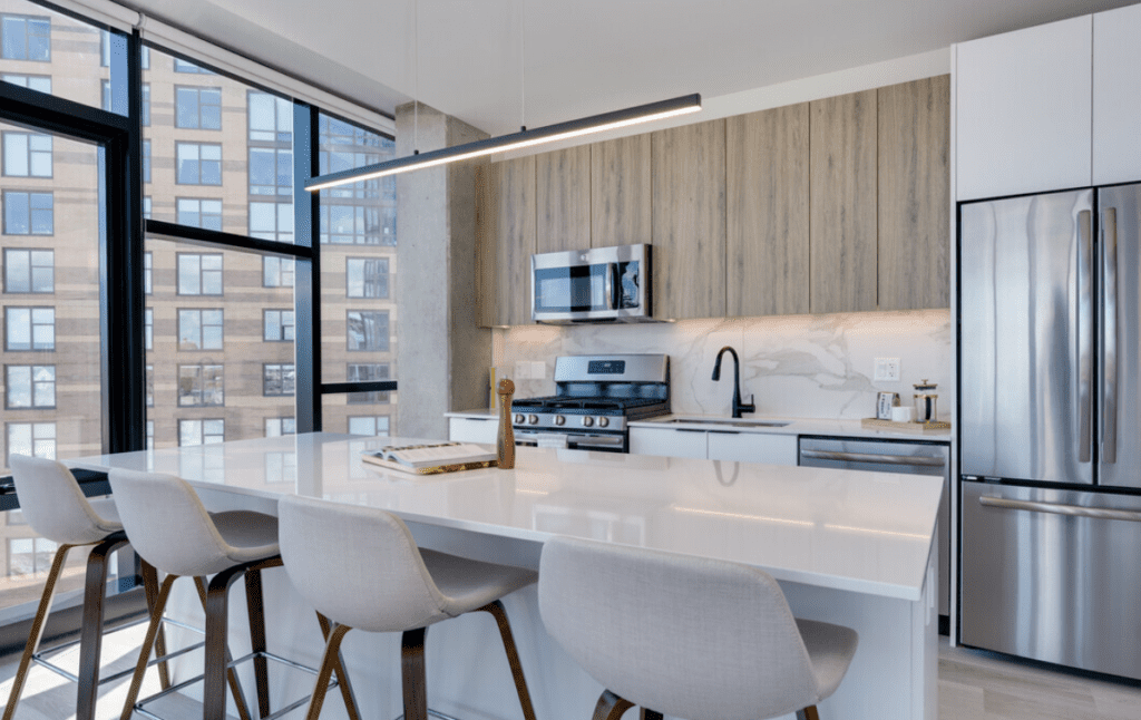 Modern kitchen with a white island, four gray stools, stainless steel appliances, and wooden cabinets. Large windows provide natural light.