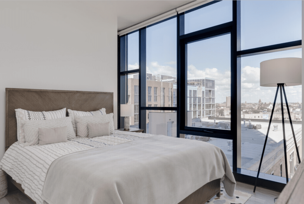 A modern bedroom with a large bed, floor-to-ceiling windows, a city view, and a tall floor lamp.