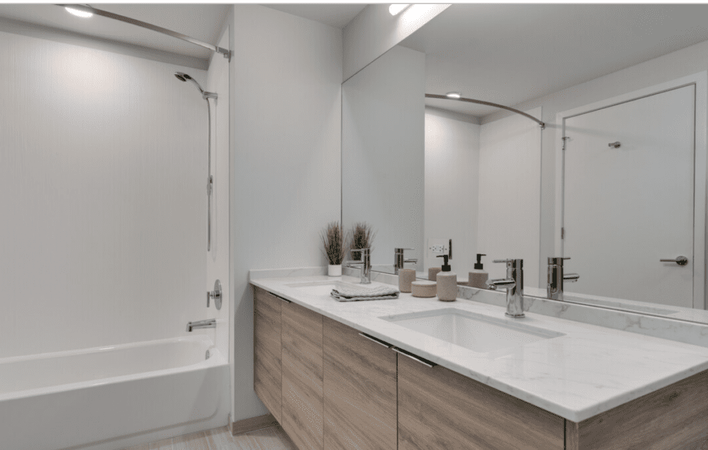 Modern bathroom with large mirror, wooden vanity with marble countertop, sink, and faucet. Tub and shower with a curved curtain rod. Minimalist decor with potted plants and toiletries.
