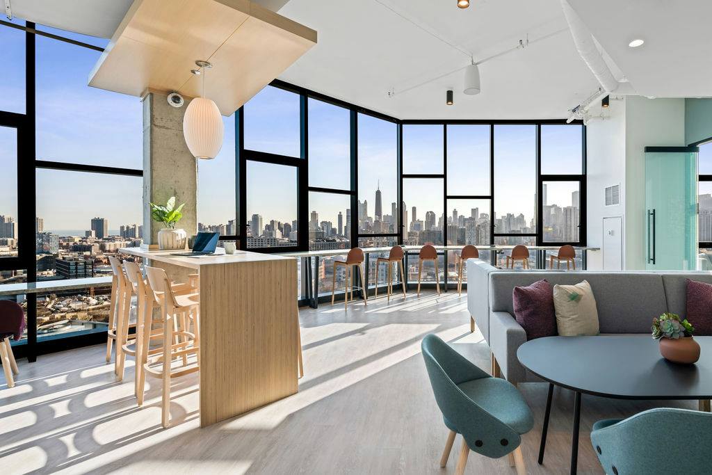 Bright modern lounge area with floor-to-ceiling windows, city skyline view, high-top tables, chairs, a sofa, and a small round table with armchairs.