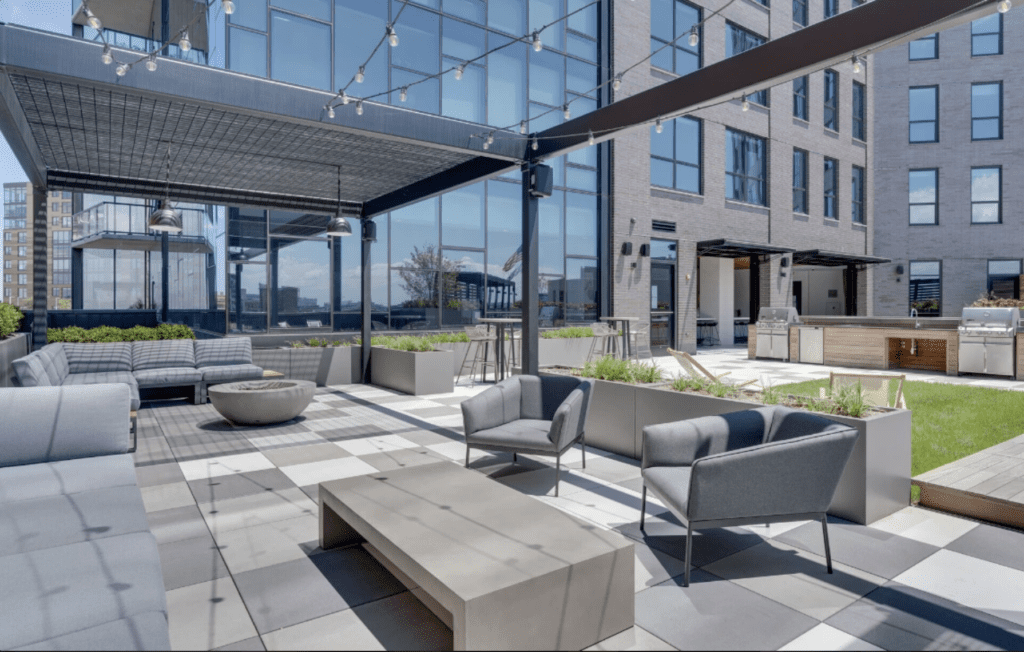 Modern rooftop patio with gray seating, a rectangular table, and a fire pit. Buildings with glass facades are in the background. String lights hang overhead.