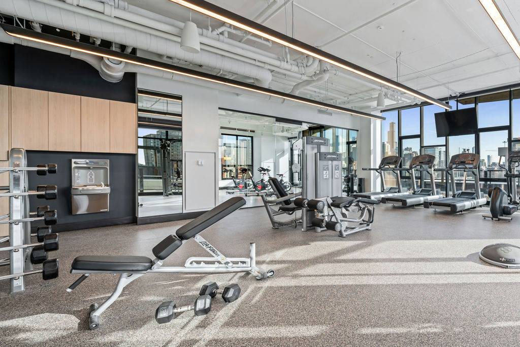 Modern gym with exercise machines, treadmills, benches, and weights in a spacious room with large windows and bright lighting.