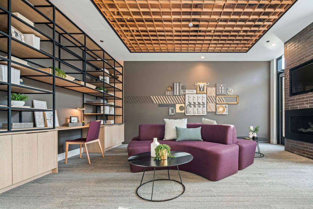 Modern room with a plush purple sofa, geometric wall decor, black metal shelving, and a wooden desk with a pink chair. A round black coffee table holds potted plants and a vase.