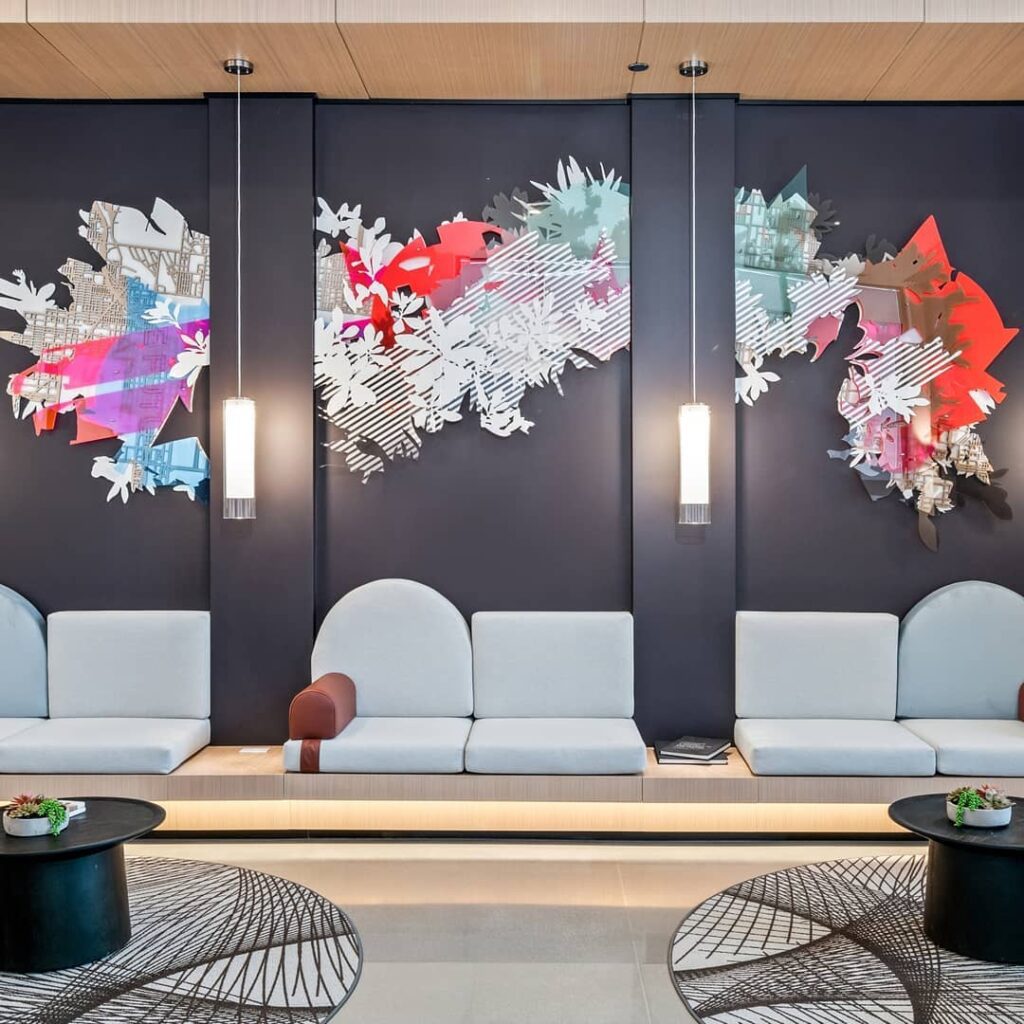 Modern lobby with minimalist seating, abstract wall art featuring colorful shapes, two hanging lights, and round black tables with small plants.