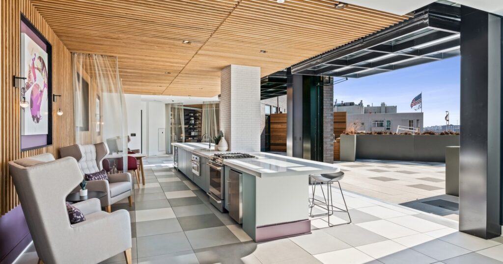 Rooftop lounge with a sleek modern kitchen, armchairs, and open-air patio. Wooden ceiling panels and a US flag in the background.