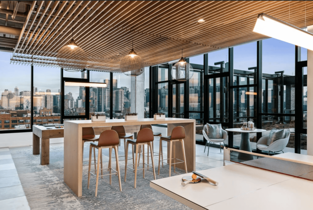 Modern rooftop lounge with high stools, a pool table, and a view of the city skyline through large windows. A wooden ceiling and hanging lights enhance the contemporary design.