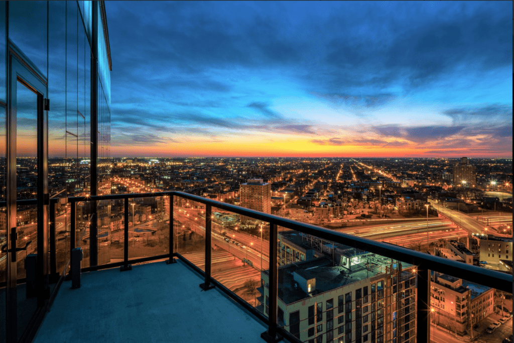 Cityscape view from a high-rise balcony at dusk, with city lights illuminating streets and a colorful sunset sky in the background.