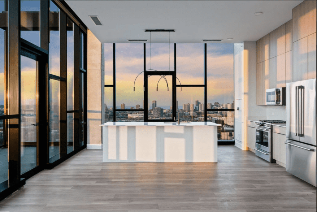 Modern kitchen with floor-to-ceiling windows, light wood flooring, and stainless steel appliances. City skyline and sunset visible through windows.