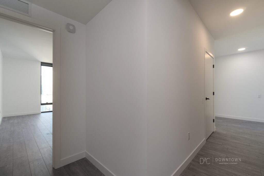 Minimalist interior of an empty apartment hallway with light wood flooring and white walls, showing a visible open doorway leading to a sunlit room.