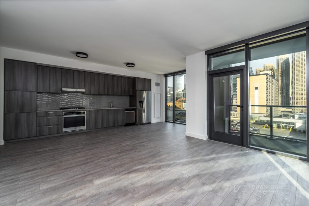 Modern, unfurnished apartment with dark wood kitchen cabinets, stainless steel appliances, light wood flooring, and large windows overlooking a cityscape.