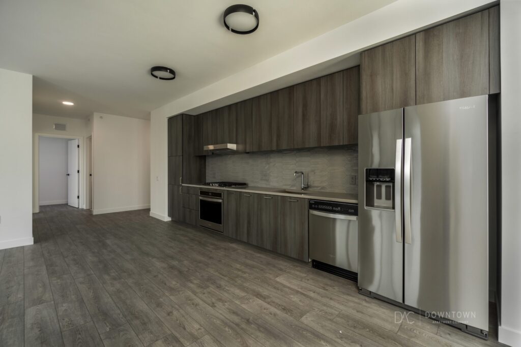 Modern kitchen with gray wood cabinets, stainless steel appliances, and wood flooring. White walls and circular ceiling lights complete the minimalist look.