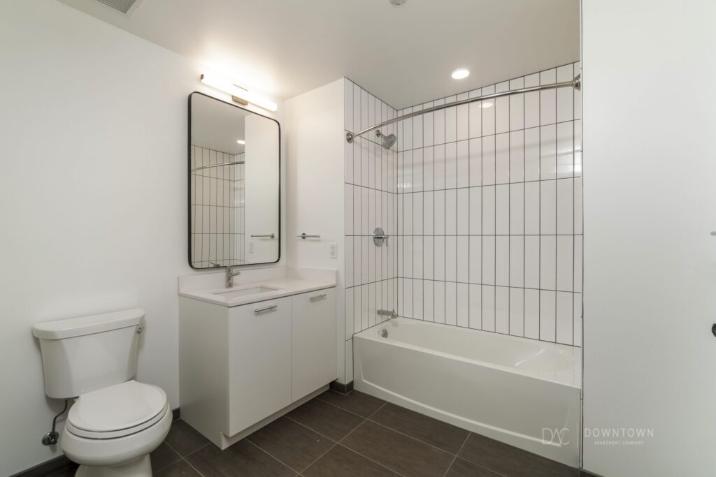 Minimalist bathroom with white walls, a bathtub with a shower, a large mirror above a white sink, and a toilet. The floor is dark tiled.