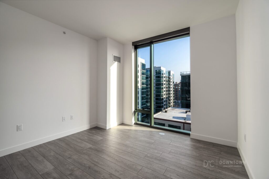 Empty room with light gray hardwood flooring and a large window providing a view of nearby buildings under a clear sky.