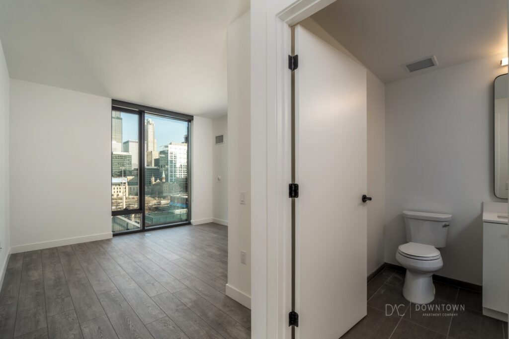 Apartment interior featuring a sunlit room with large windows and wood flooring on the left, adjacent to a bathroom with a toilet and sink on the right.