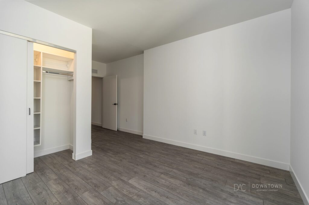 Empty room with white walls, gray wood flooring, open closet with sliding door, and a closed door. Natural light illuminates the space.