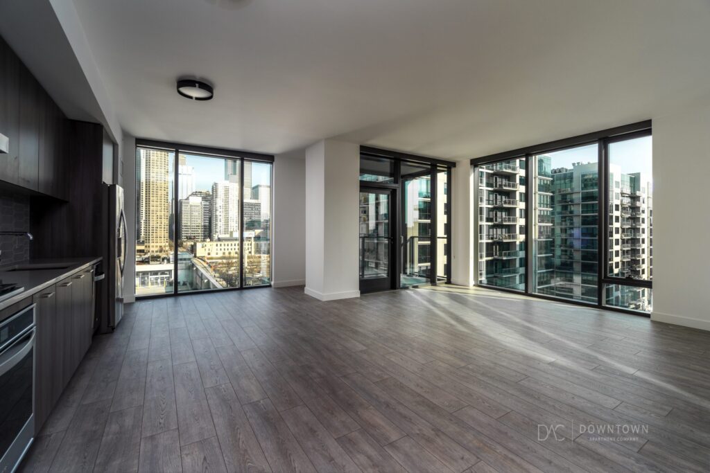 Modern apartment interior with large windows, city view, dark wood flooring, and a kitchen on the left.