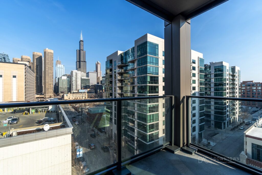 View from a balcony overlooking modern high-rise buildings and the city skyline under a clear blue sky. The Willis Tower is visible in the background.
