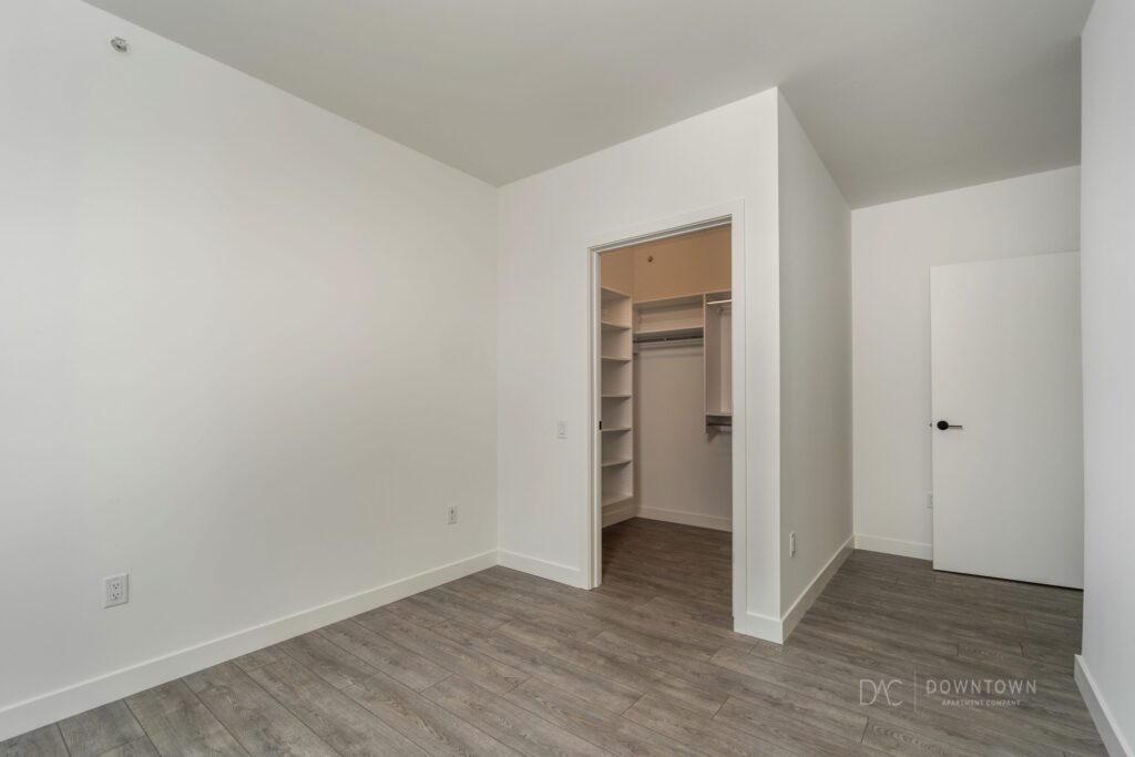 An empty room with light gray walls and wood flooring, featuring an open door to a small closet with shelves and hanging space.
