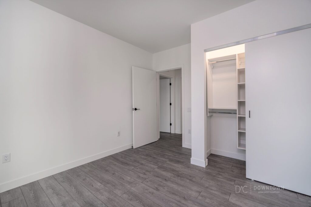 Empty room with gray wood flooring, white walls, an open closet with shelves, and two doors, one leading to a hallway.