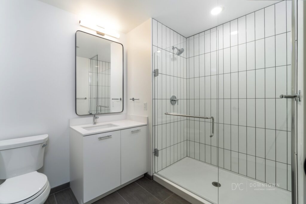 Modern bathroom with white vanity, rectangular mirror, and walk-in glass shower featuring vertical white tiles. Brown tiled floor, with toilet next to the vanity.
