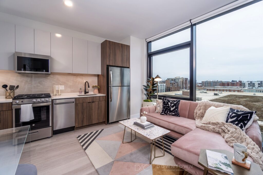 Modern apartment living room and kitchen with a pink sectional sofa, geometric rug, glass coffee table, stainless steel appliances, and large windows overlooking a cityscape.