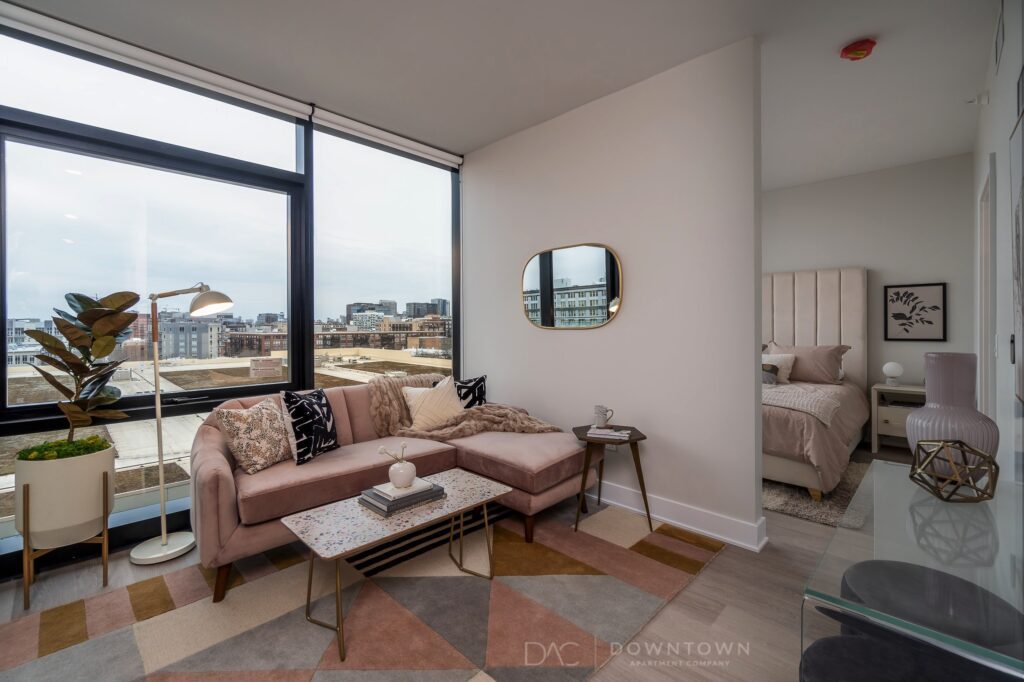 Modern apartment with large windows, a beige sectional sofa, geometric rug, and decorative items. A separate bedroom area is visible with a bed and side tables. Cityscape view outside.