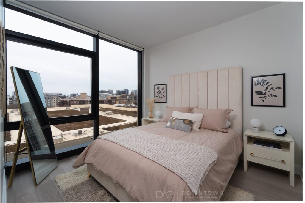 Bedroom with large window, city view, bed with beige linens, a tall mirror, nightstands with lamps, and wall art.