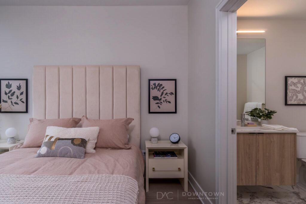 Bedroom with a beige bed, pink bedding, and two nightstands. Decor includes leafy artwork above the bed. An adjacent bathroom has a vanity and mirror with soft lighting.