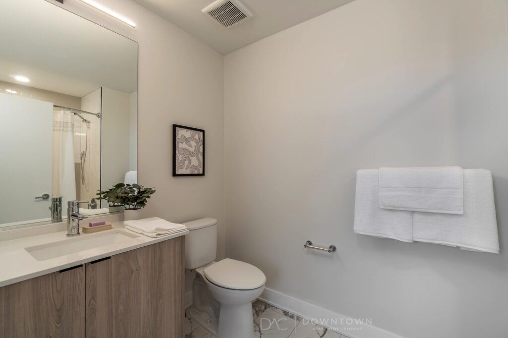 Modern bathroom with a toilet, wooden vanity with sink, mirror, potted plant, wall art, towel rack with white towels, and a shower area with a curtain.
