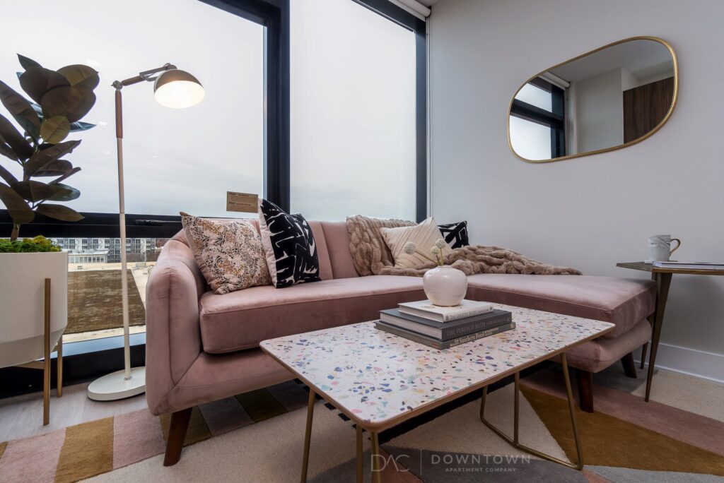 A modern living room features a pink sofa with patterned cushions, a terrazzo coffee table, a lamp, and a mirror on the wall. Large windows offer a city view.