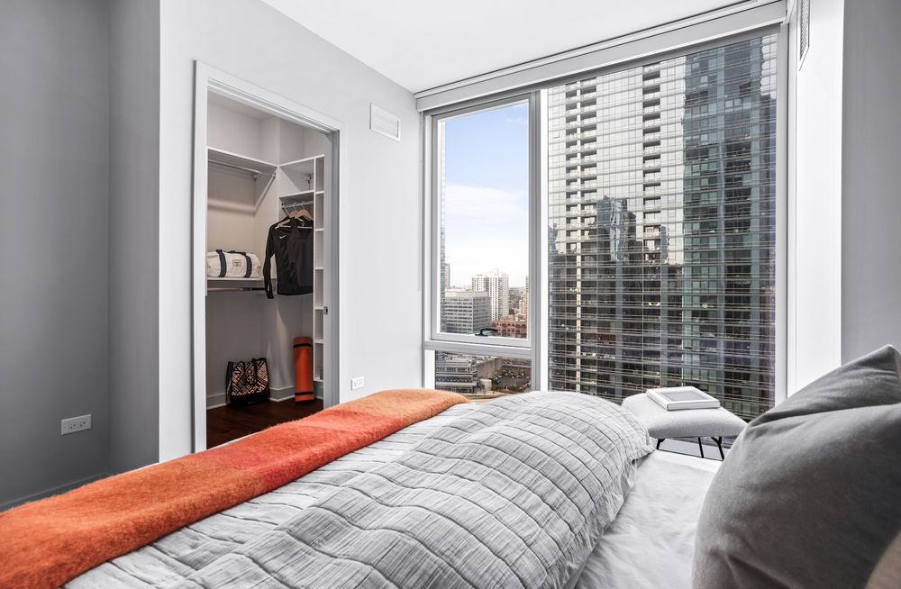 Modern bedroom with a neatly made bed, adjacent walk-in closet, and a large window offering a cityscape view of tall buildings.