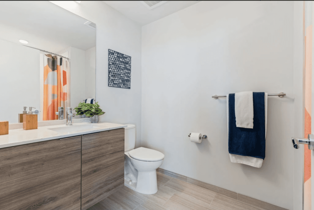 Modern bathroom with a wooden vanity, white countertop, potted plant, toilet, wall-mounted towel rack with blue and white towels, and a shower with a colorful curtain.