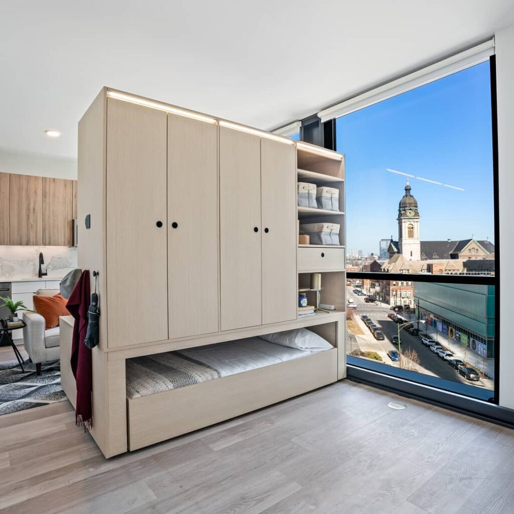 Compact, modern apartment interior featuring a multifunctional wooden cabinet unit with storage and a bed, next to a large window overlooking a cityscape with a historic building.