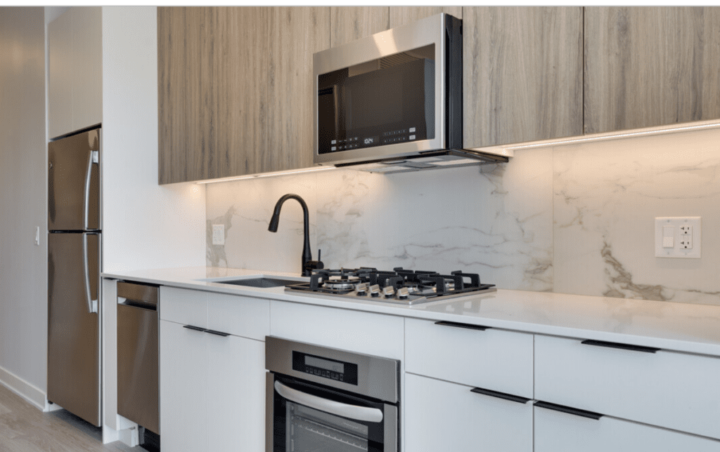 Modern kitchen with stainless steel appliances, including a refrigerator, dishwasher, and oven, with wood and white cabinetry, marble backsplash, and a black faucet.