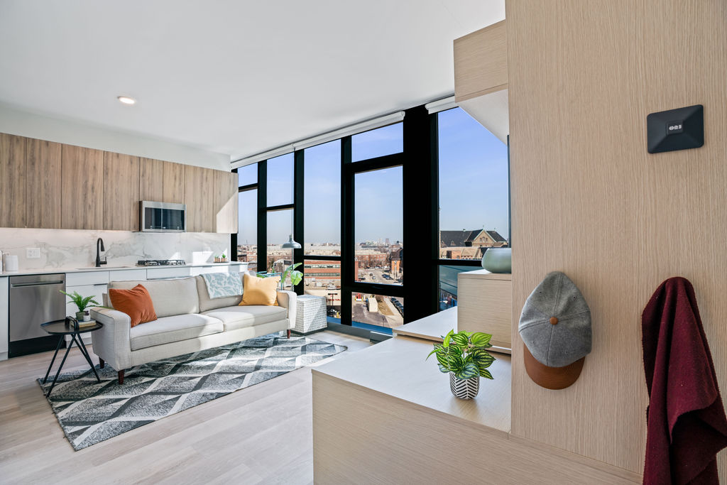 Modern living room with large windows, a light gray sofa, and a geometric rug. The kitchen area features wood cabinets and a marble backsplash. A hat and jacket hang on a wall in the foreground.