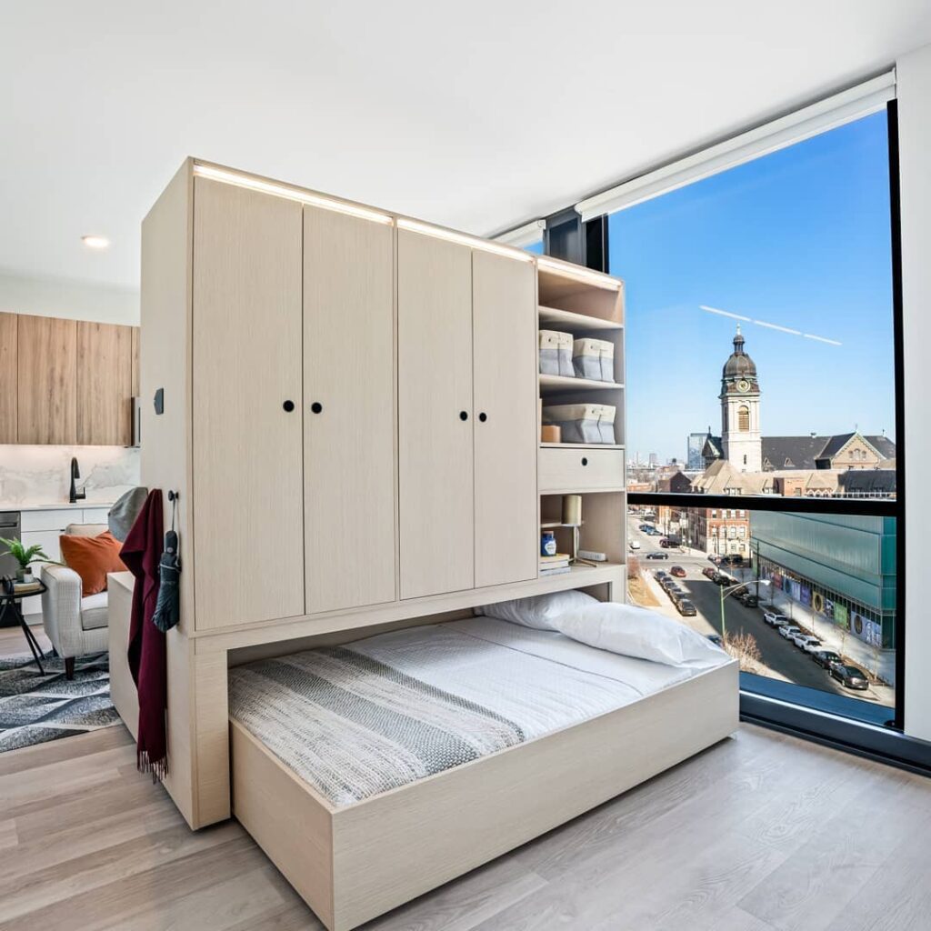 Compact apartment with a pull-out bed and built-in shelves, overlooking a cityscape with a large church and modern buildings visible through the window.