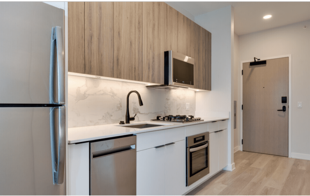 Modern kitchen with stainless steel appliances, marble backsplash, and light wood cabinets. Features a refrigerator, dishwasher, oven, microwave, and a black faucet. Light wood flooring.