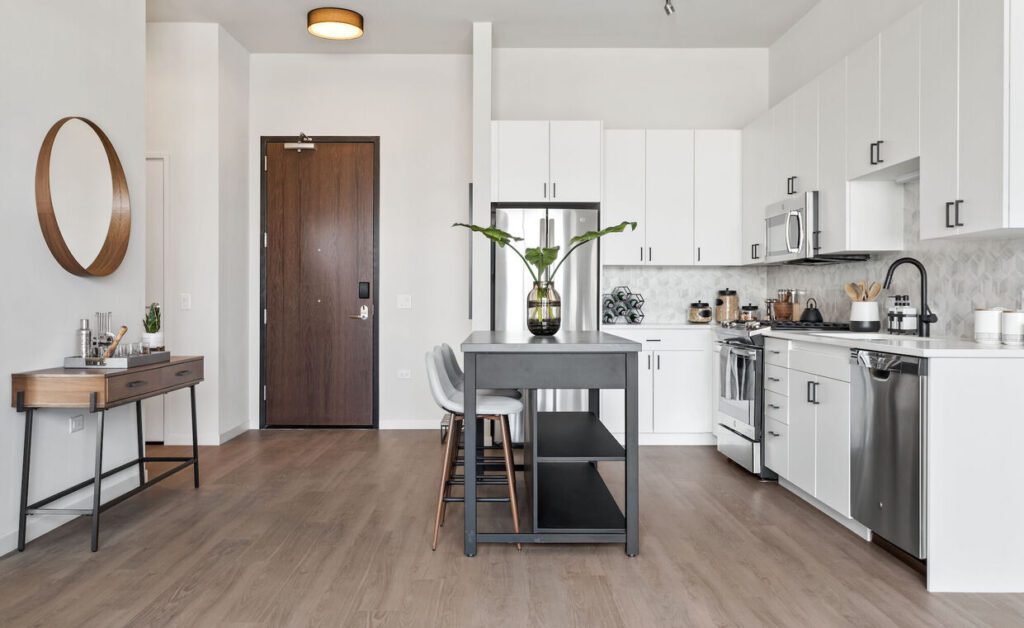 Modern kitchen with white cabinets, stainless steel appliances, and a central gray island. A round mirror and console table decorate the entryway to the left, with light wood flooring throughout.