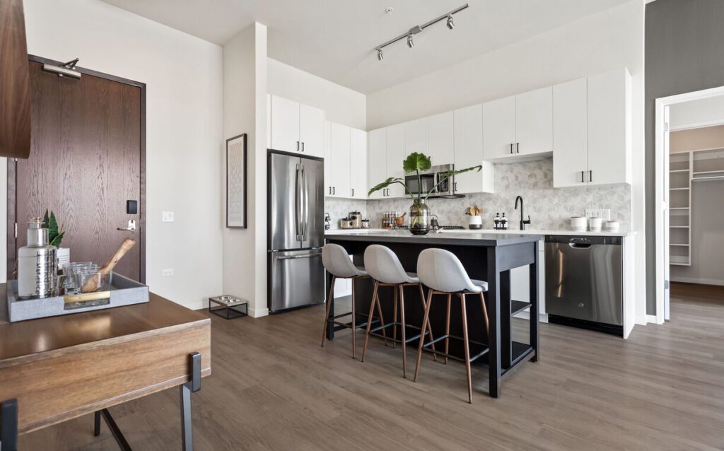 Modern kitchen with white cabinets, stainless steel appliances, and a dark island with three gray bar stools. Light wood flooring and a decorative plant on the island.