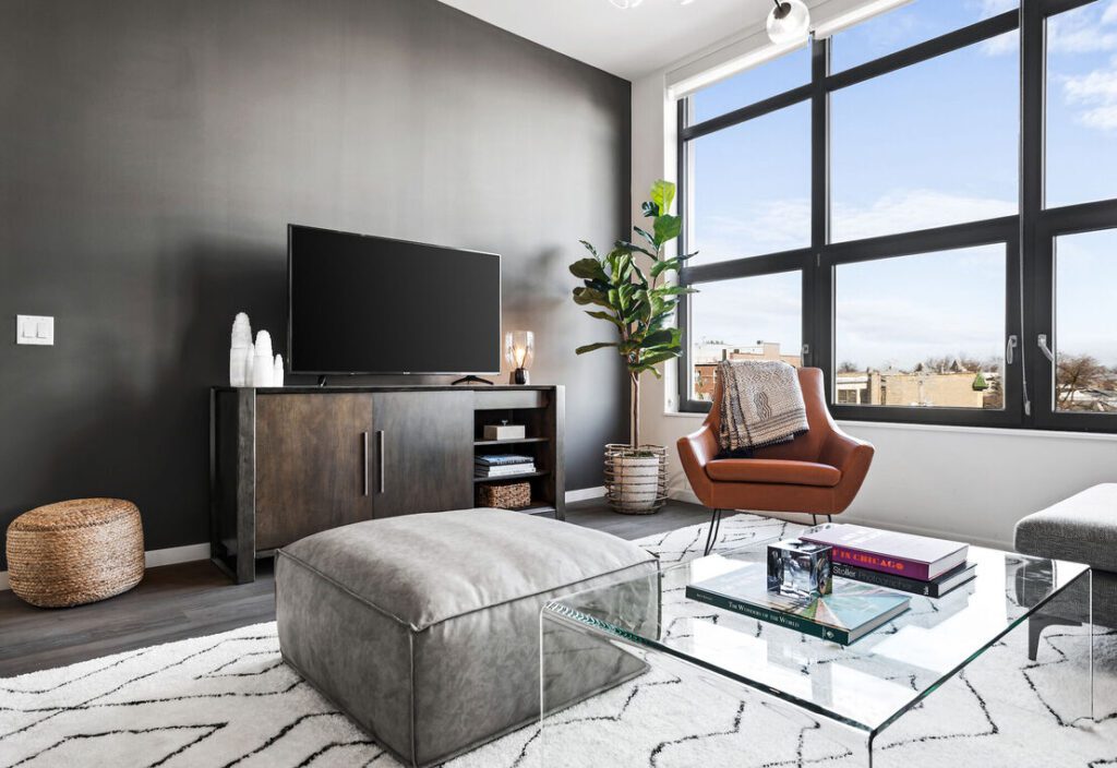 Modern living room with a dark accent wall, TV on a console, leather chair, large window, plant, and glass coffee table on a patterned rug.