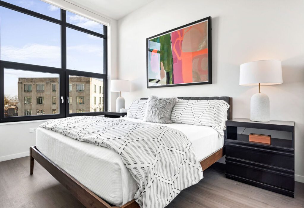 Modern bedroom with a large window, abstract art, and double bed with patterned bedding. Two lamps rest on dark bedside tables with a view of a building outside.