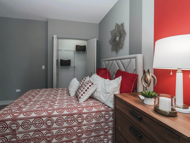 Bedroom with a bed featuring red and white patterned bedding, a nightstand with a lamp and decor, and an open closet with shelves. Room has gray and red walls.