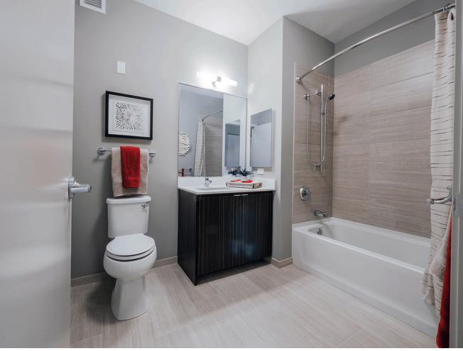 Modern bathroom with a bathtub and shower, toilet, sink with mirror, artwork, and red towels. Neutral-colored tiles and gray walls enhance the clean, minimalist design.